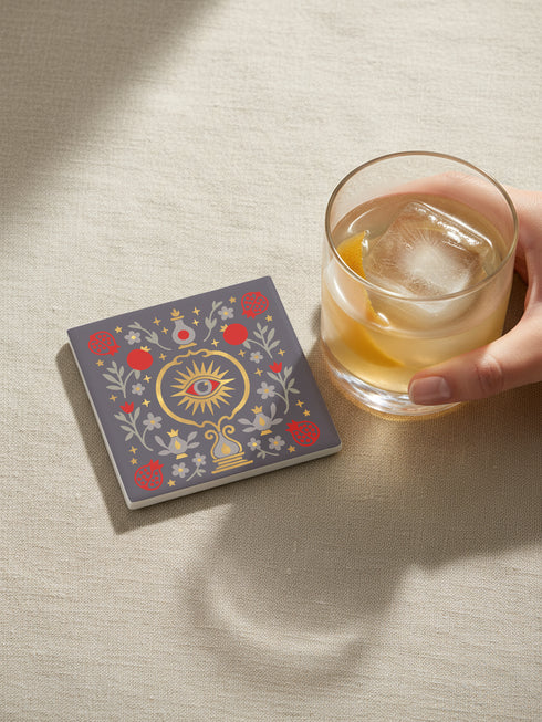 A square ceramic drink coaster with a smooth, matte charcoal grey finish. The surface features a symmetrical, intricate design in metallic gold, deep garnet red, and light grey. In the center is a golden All-Seeing Eye with a red iris, radiating light. The eye is framed by a golden circle and flanked by botanical elements, including red pomegranates and grey flowers with gold details. The design has a folk art, gothic botanical style.